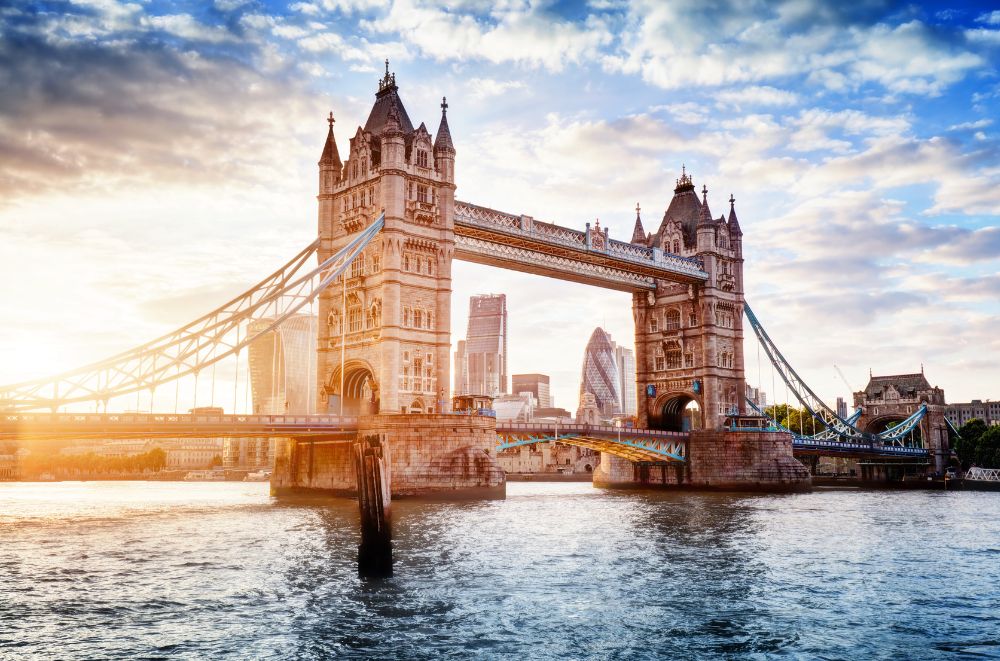 The iconic Tower Bridge in London spans the River Thames under a partly cloudy sky. The sun is setting, casting a warm, golden light on the bridge and the water. The modern skyline, including the distinctive Gherkin building, is visible in the background.