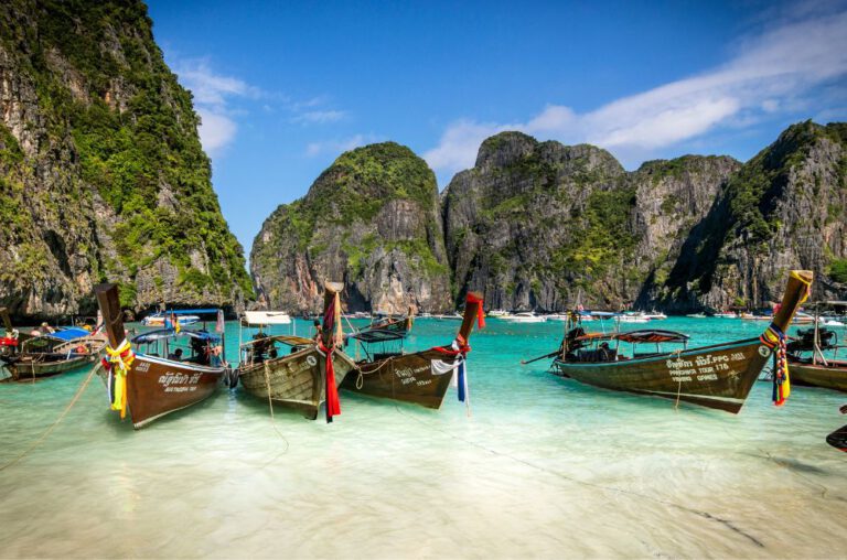 Colorful traditional Thai boats moored in clear blue water, with lush green mountains and a bright sky in the background.