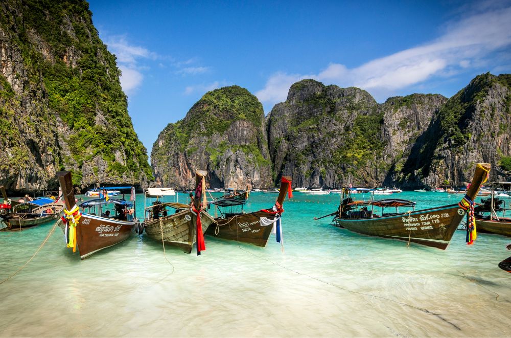 Colorful traditional Thai boats moored in clear blue water, with lush green mountains and a bright sky in the background.