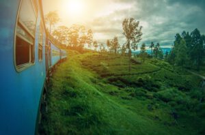 A blue train curves along a lush, green hillside under a cloudy sky in Sri Lanka. The landscape is dotted with trees and vegetation, and the sun is shining through the clouds, casting a warm glow over the scene.