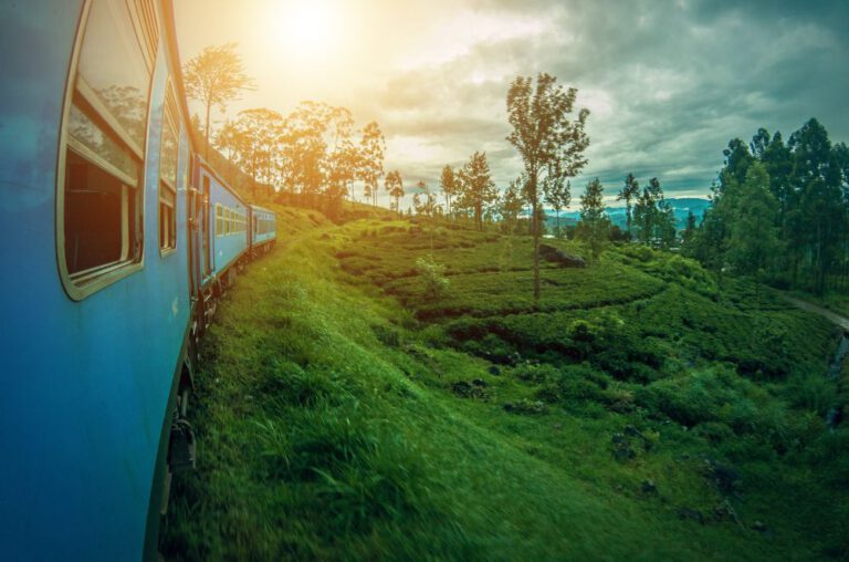 A blue train curves along a lush, green hillside under a cloudy sky in Sri Lanka. The landscape is dotted with trees and vegetation, and the sun is shining through the clouds, casting a warm glow over the scene.
