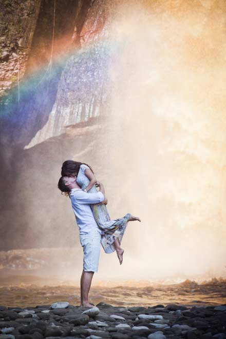 A couple embraces joyfully as the man lifts the woman off the ground, standing on rocky terrain with a gentle rainbow and misty waterfall in the background.