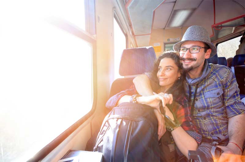 A smiling couple sits close together on a train, looking out the window at the bright sunlight. The woman leans on the man, and both appear happy and relaxed, enjoying their journey.