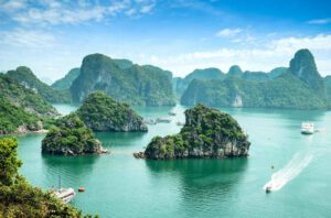 A scenic view of Ha Long Bay featuring emerald-green water, several lush, rocky islands, boats cruising, and a backdrop of misty limestone karsts under a blue sky with scattered clouds.
