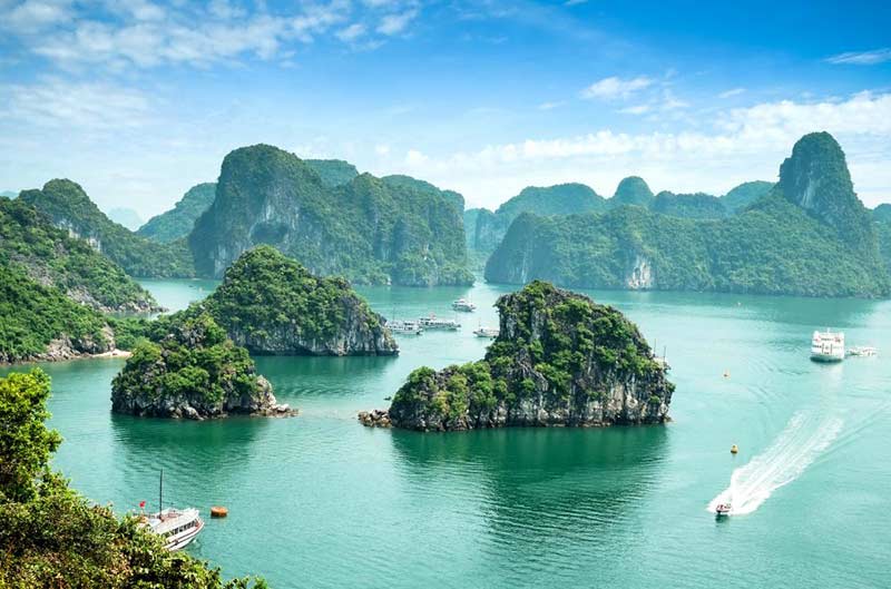 A scenic view of Ha Long Bay featuring emerald-green water, several lush, rocky islands, boats cruising, and a backdrop of misty limestone karsts under a blue sky with scattered clouds.