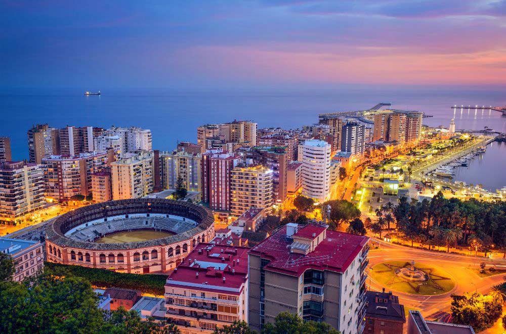 Aerial view of Málaga, Spain at sunset, showing the illuminated city, a large round bullring, tall buildings, and the coastline with the sea in the background.