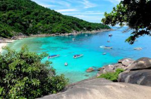 A tropical bay with turquoise water, several boats floating near the shore, lush green hills in the background, and rocks and foliage in the foreground under a blue sky.