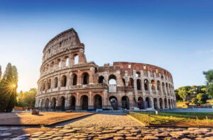 The Colosseum in Rome, Italy, illuminated by sunlight under a clear blue sky, with ancient stone arches and ruins visible in the foreground.