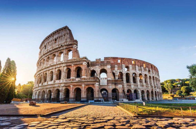 The Colosseum in Rome, Italy, illuminated by sunlight under a clear blue sky, with ancient stone arches and ruins visible in the foreground.