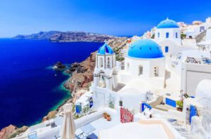 White buildings with blue domes overlook the deep blue sea and rocky coastline under a clear sky in Santorini, Greece. The architecture stands out against the vibrant ocean and distant cliffs.
