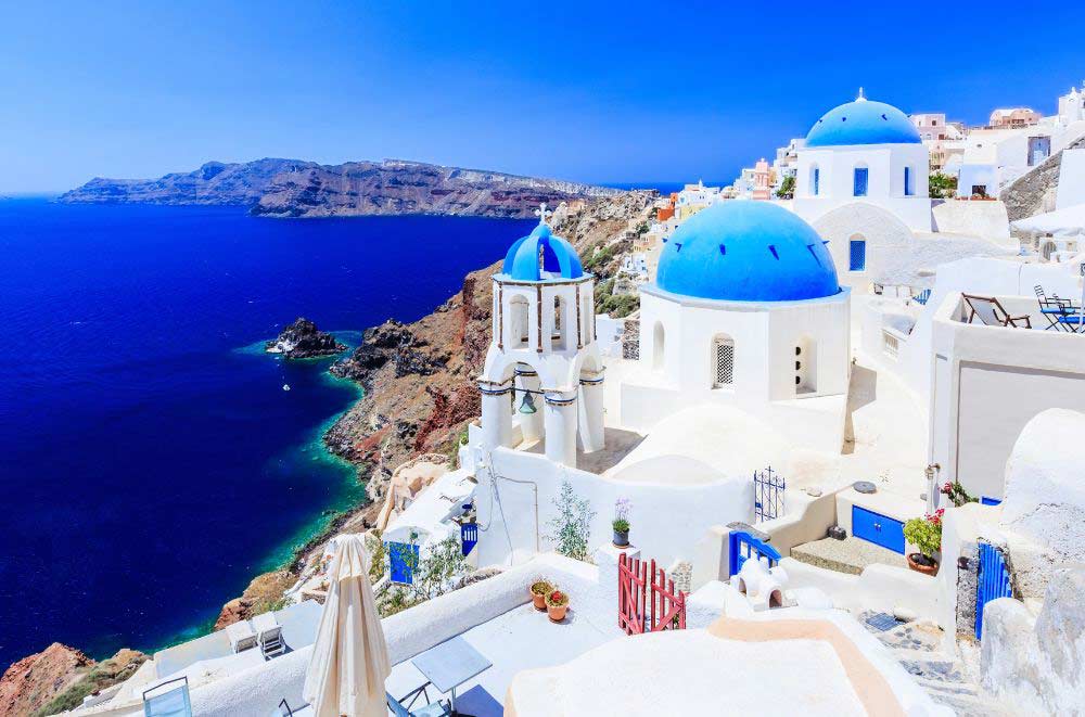White buildings with blue domes overlook the deep blue sea and rocky coastline under a clear sky in Santorini, Greece. The architecture stands out against the vibrant ocean and distant cliffs.