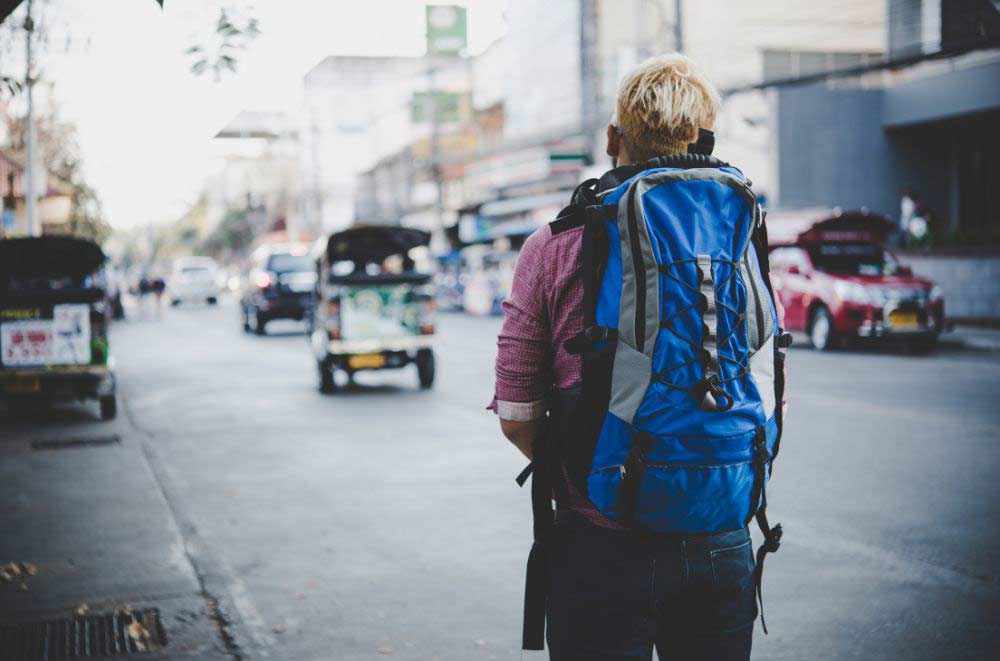 A person with blond hair and a blue backpack stands on a city street, facing away from the camera, as cars and tuk-tuks drive by in the background.