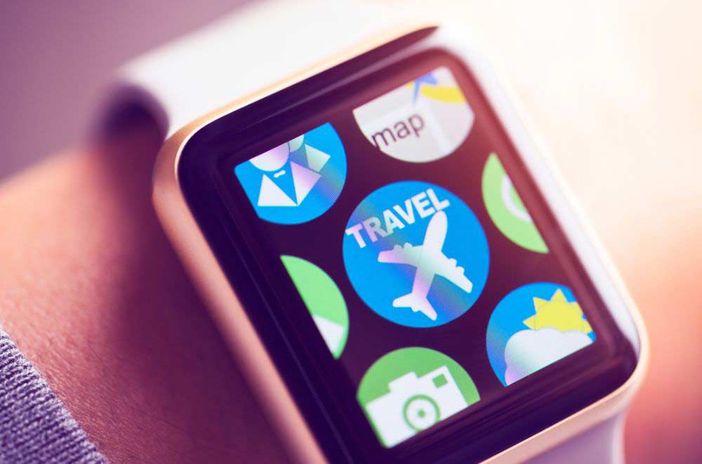 A close-up of a smartwatch screen displaying colorful app icons, including a travel app with an airplane, a camera app, a map, and weather, on a persons wrist.