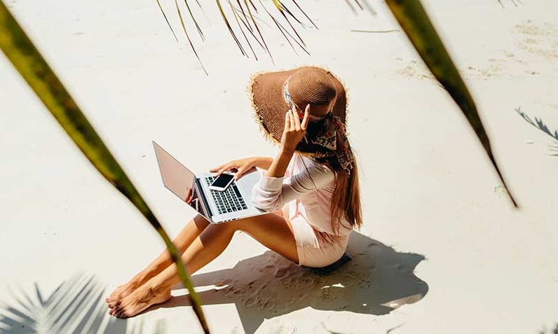 A woman in a sun hat sits on the sandy beach, using a laptop and holding a smartphone. Palm leaves frame the scene, and she is surrounded by sunlight.